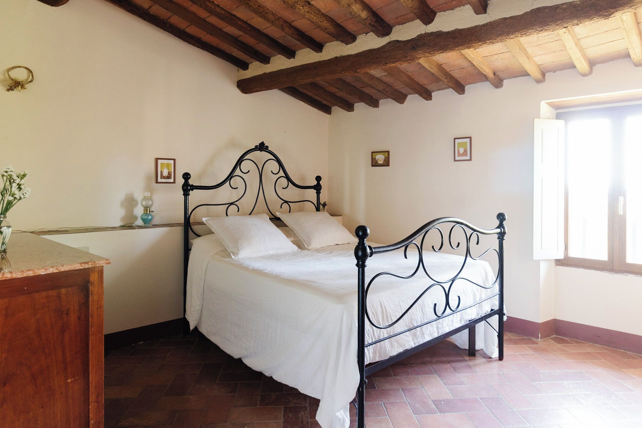 A bedroom with a bed, a nightstand, a vase with flowers, and a window.