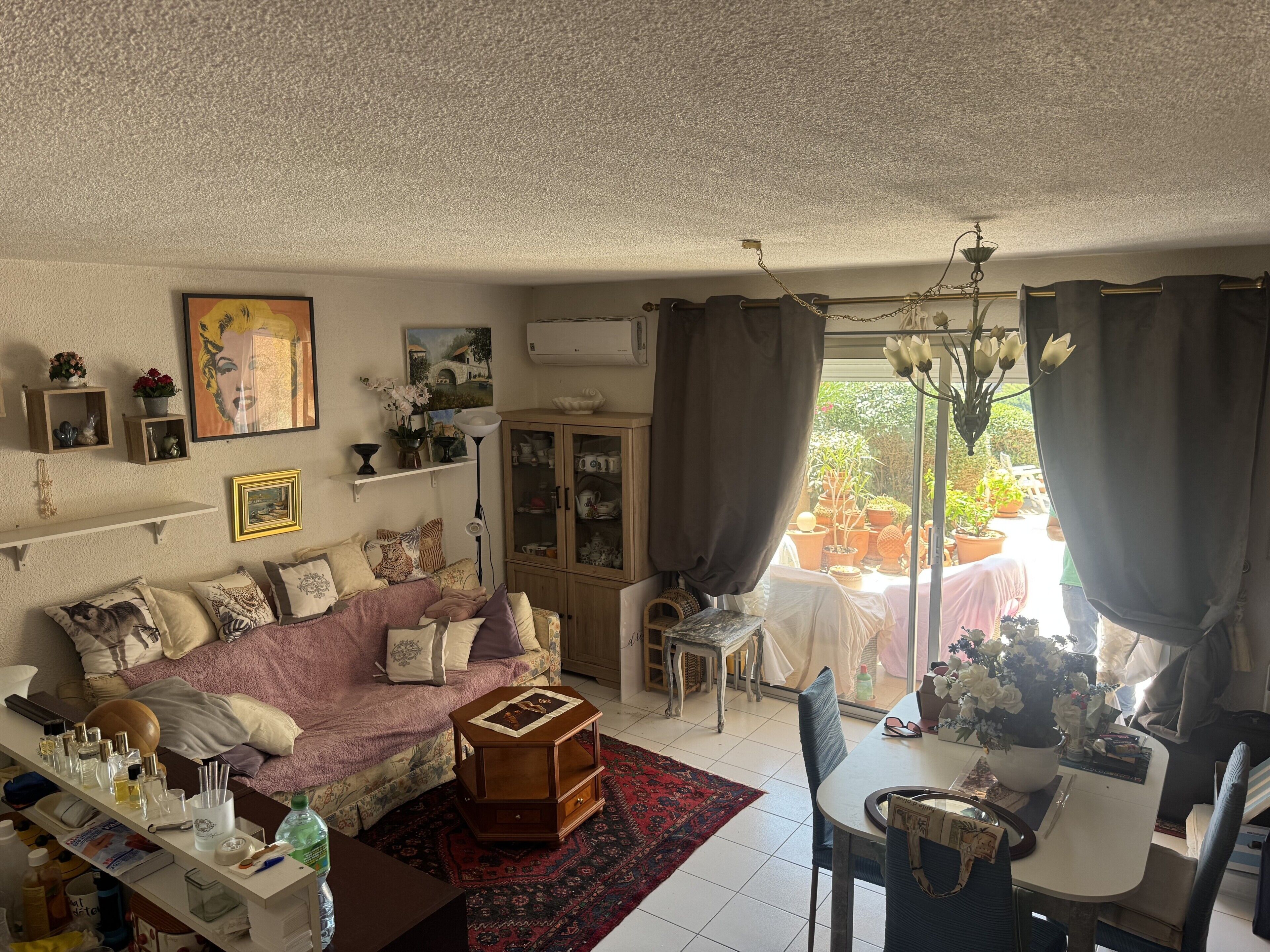 A cozy living room with a sofa, a small table, and a cabinet. There is a window with curtains, and a dining area with a table and chairs.