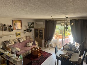 A cozy living room with a sofa, a small table, and a cabinet. There is a window with curtains, and a dining area with a table and chairs. - La Marianne with Sea View, Private Terrace, Air Conditioning (Fleury)