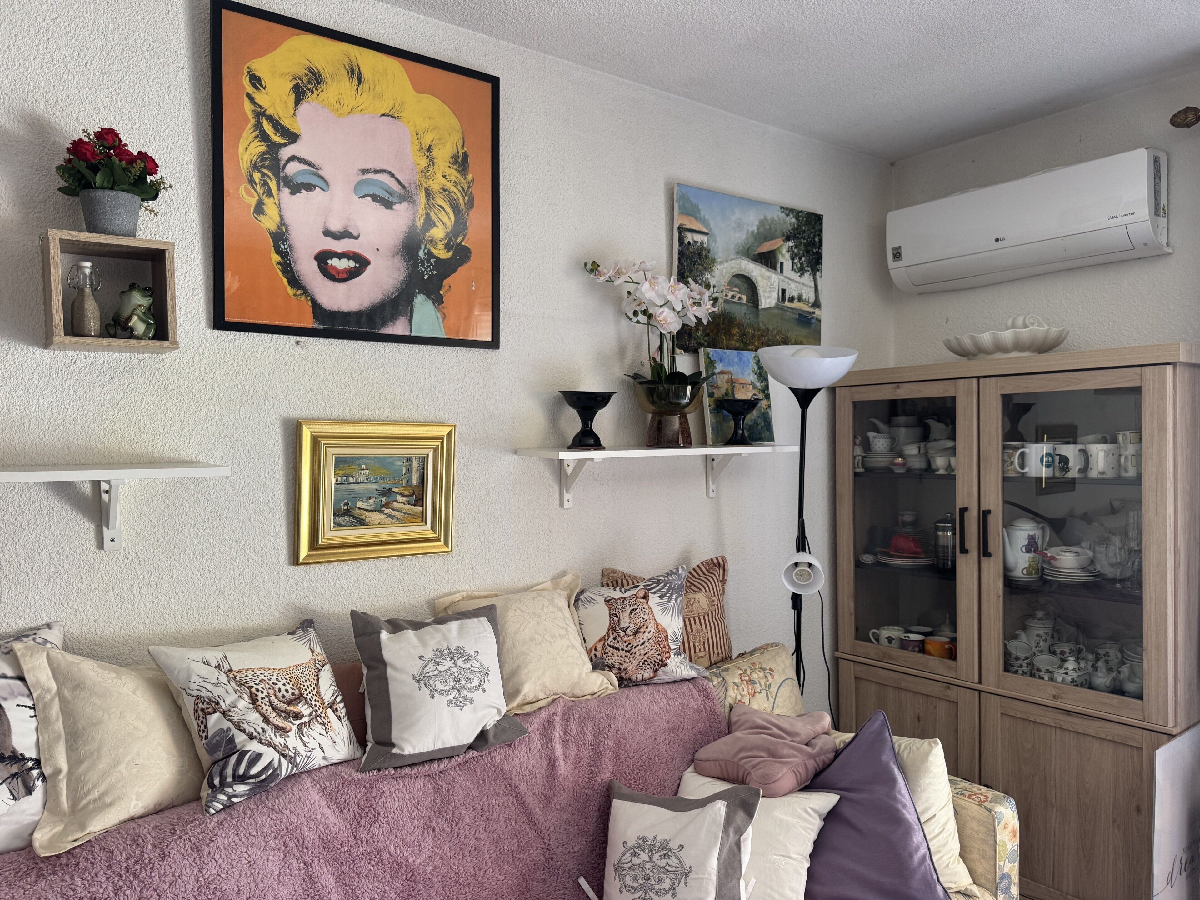 En stue med sofa, puder, et billede, en vase med blomster, et skab med service og en klimaanlæg.