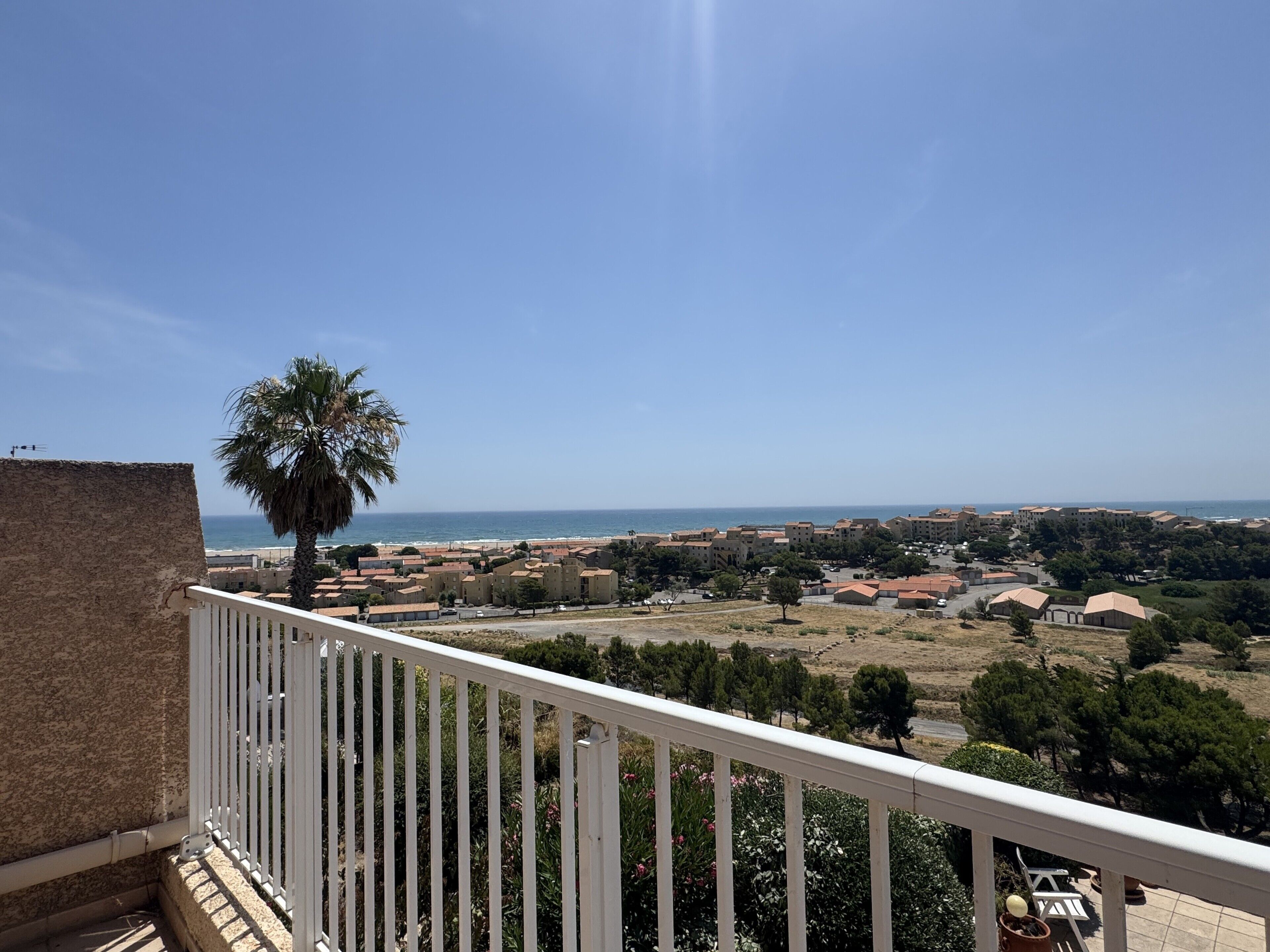 A balcony with a view of a coastal town, including buildings and a palm tree.