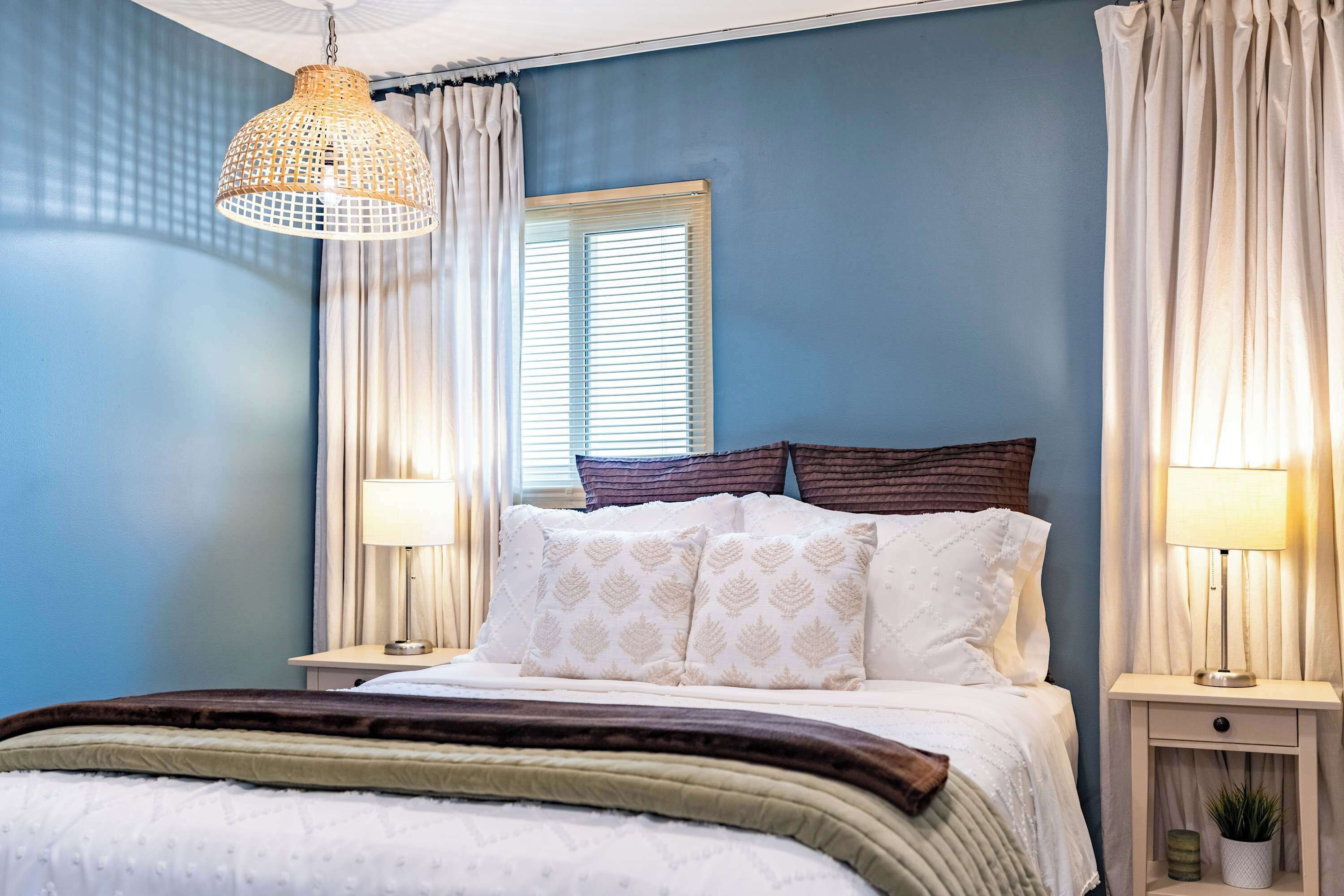 Une chambre à coucher comprenant un lit, deux tables de chevet, un luminaire suspendu et une fenêtre avec des stores.