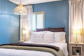 Une chambre à coucher comprenant un lit, deux tables de chevet, un luminaire suspendu et une fenêtre avec des stores.