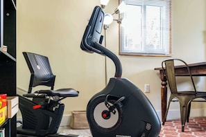 Un vélo d’appartement, une console de commande, un fauteuil de bureau noir, une table à manger et une fenêtre avec des stores.
