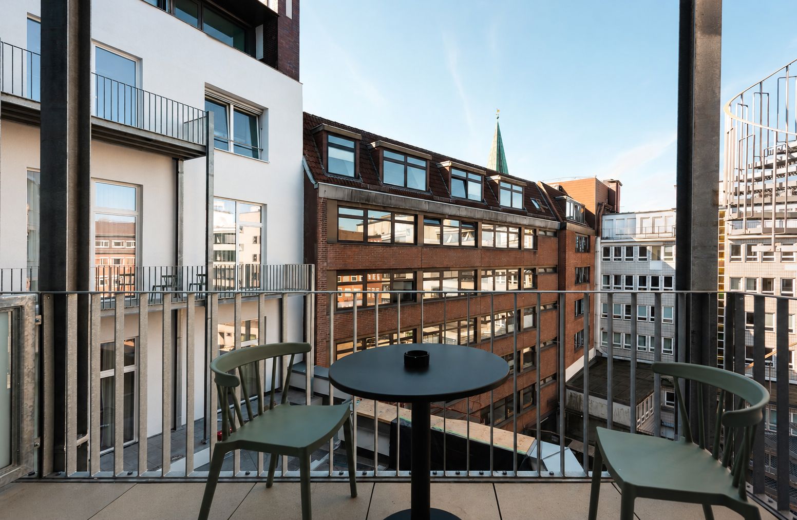 Un balcon avec une table ronde et deux chaises, d’où l’on peut admirer un immeuble à plusieurs étages, doté d’une façade en briques et d’une flèche verte.