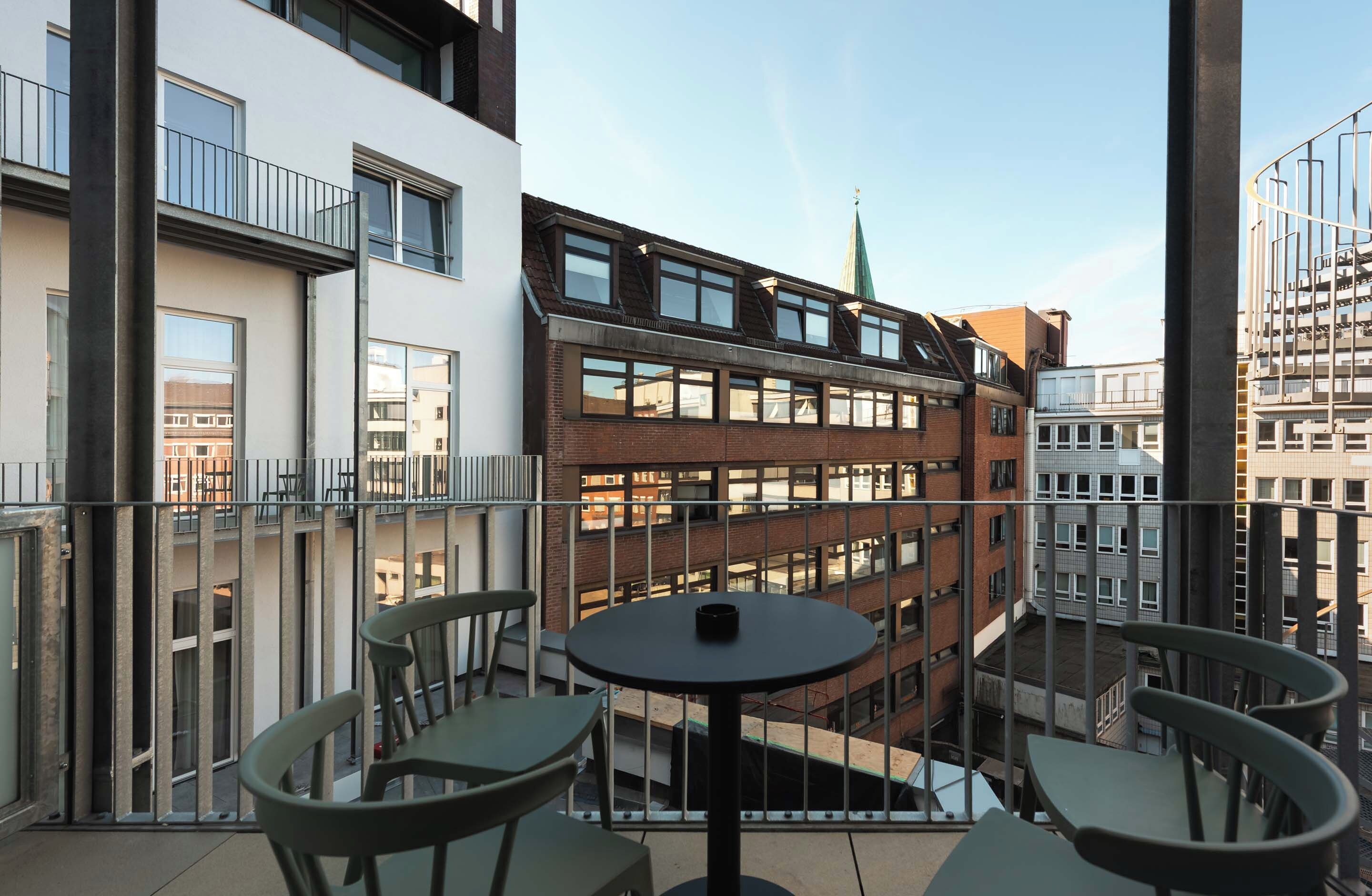En balkon med et bord og stole, med udsigt over en bygning i flere etager med en blanding af mursten og moderne arkitektur.