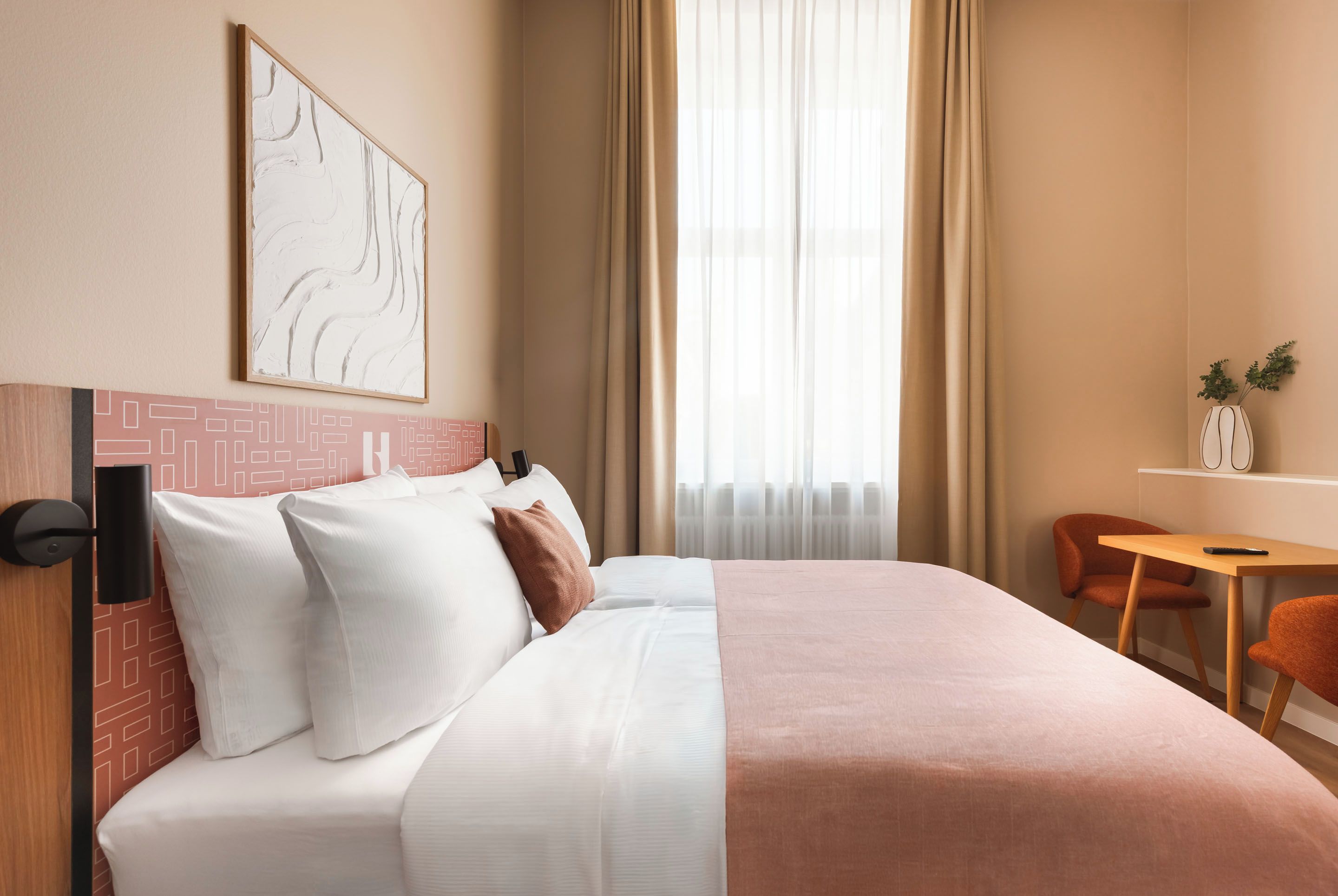 Een hotelkamer met een bed, een bureau, een stoel, een raam met gordijnen en een kunstwerk aan de muur.