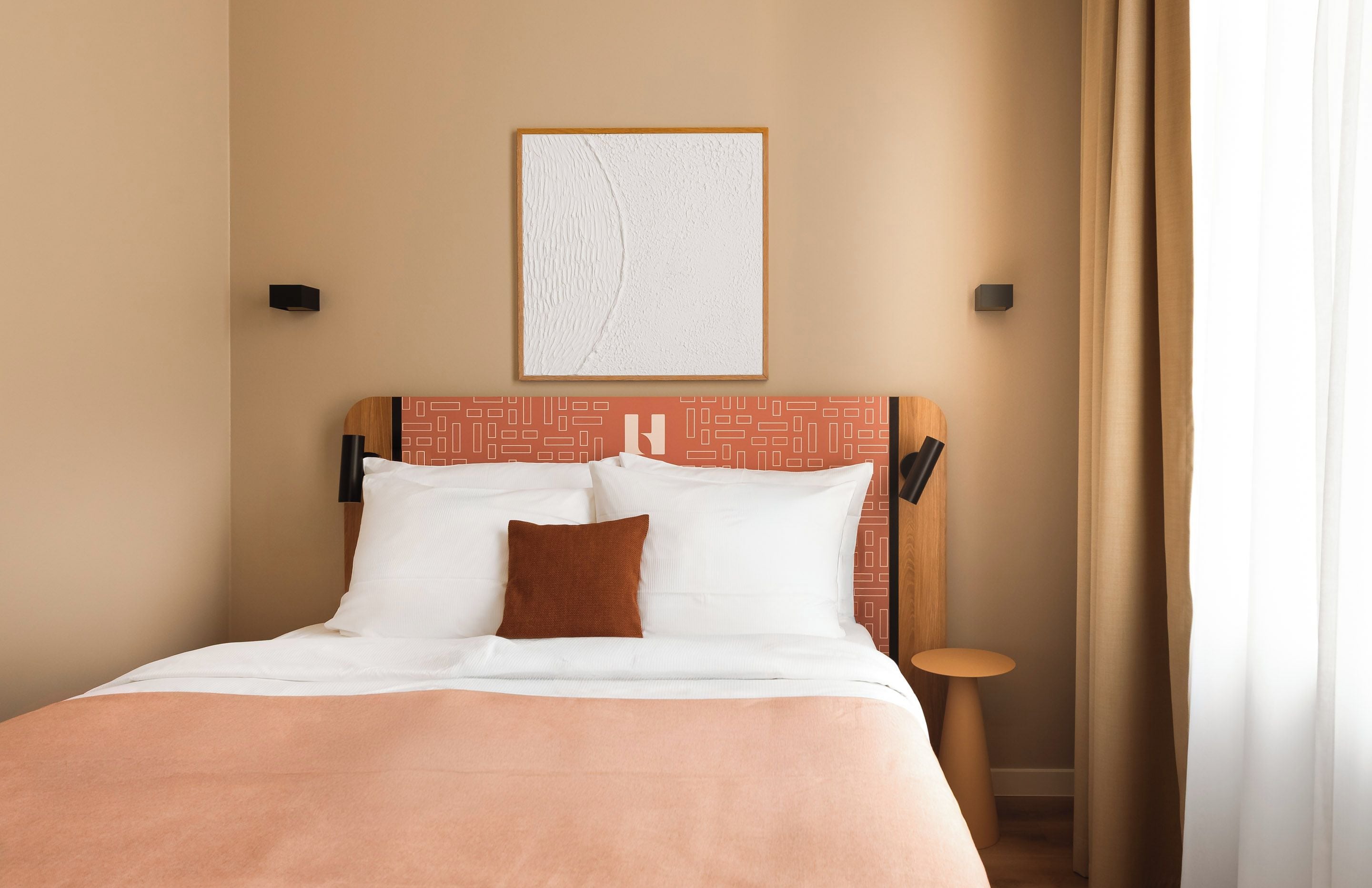 Een hotelkamer met een bed, een hoofdbord met een geometrisch patroon, een kunstwerk in een lijst aan de muur en een klein tafeltje.