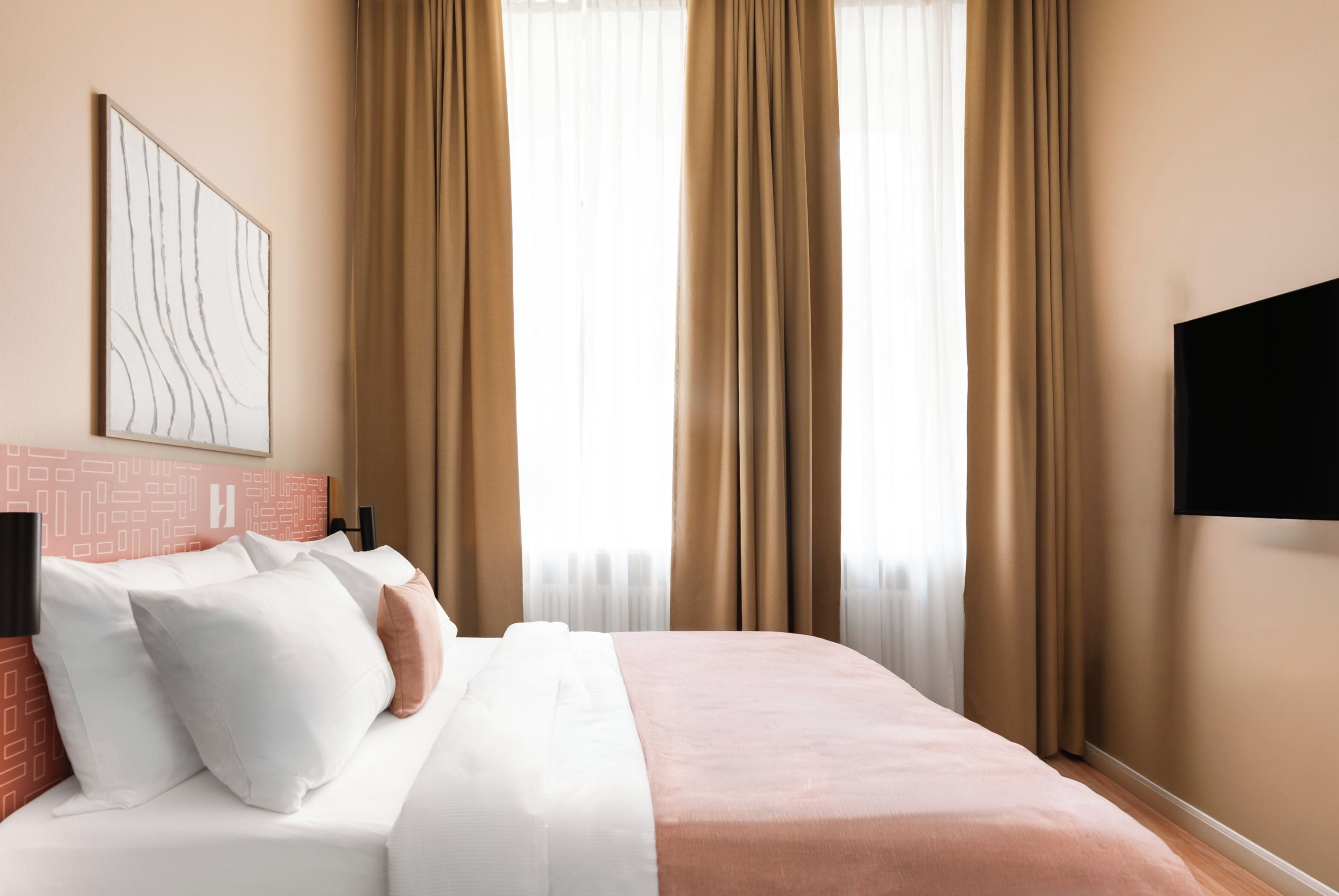 Habitación de hotel con cama, almohadas, televisión montada en la pared y ventana con cortinas.
