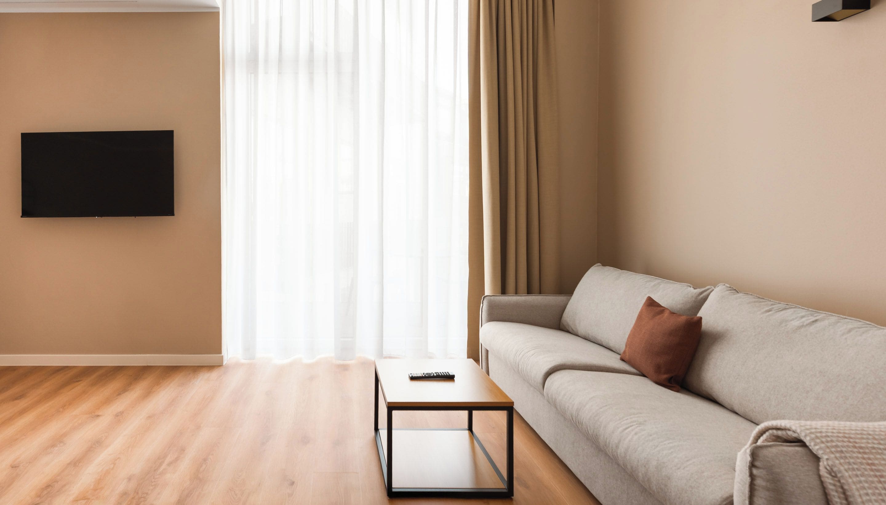 Una sala de estar con sofá, mesa de centro, televisor de pantalla plana montado en la pared y ventana con cortinas traslúcidas.