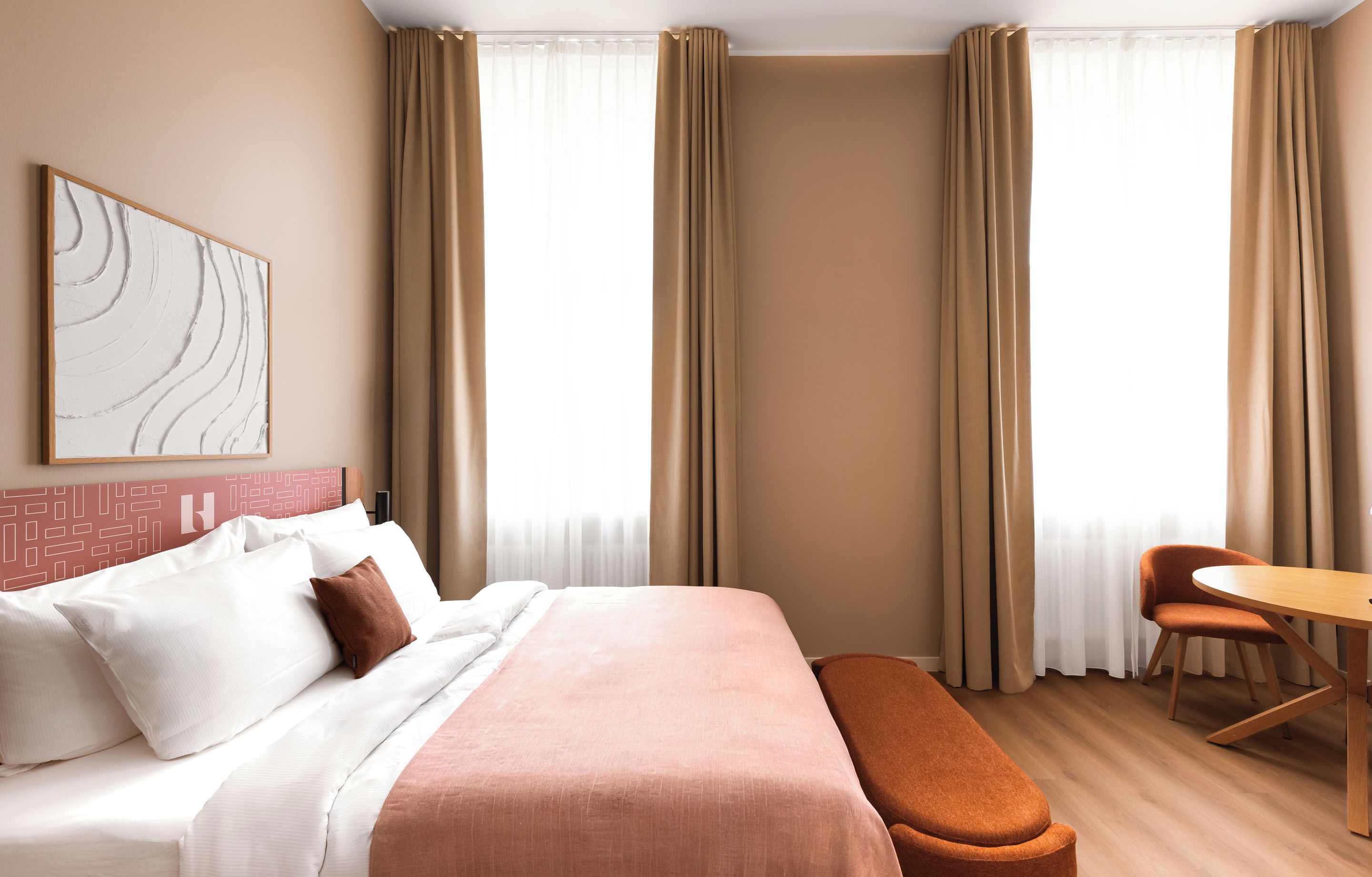 Ein Hotelzimmer mit Bett, Schreibtisch und Stuhl. An der Wand hängt ein Bild und es gibt ein Fenster mit Vorhängen.