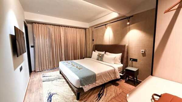 A hotel room with a large bed, a bedside table, a TV mounted on the wall, and a window with curtains.