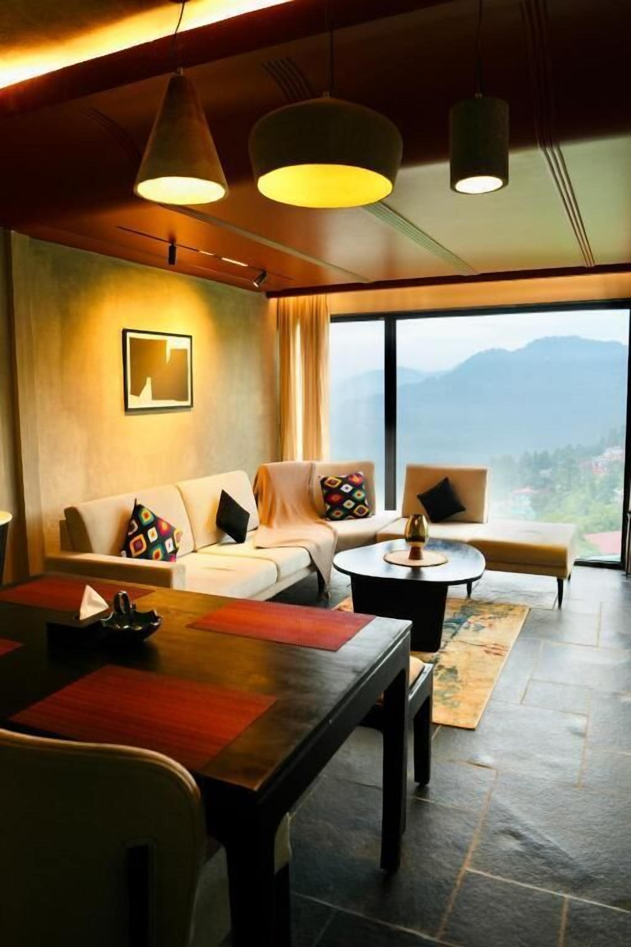 Habitación de hotel moderna con un conjunto de sofás, una mesa de comedor y vistas a montañas a través de ventanas amplias.