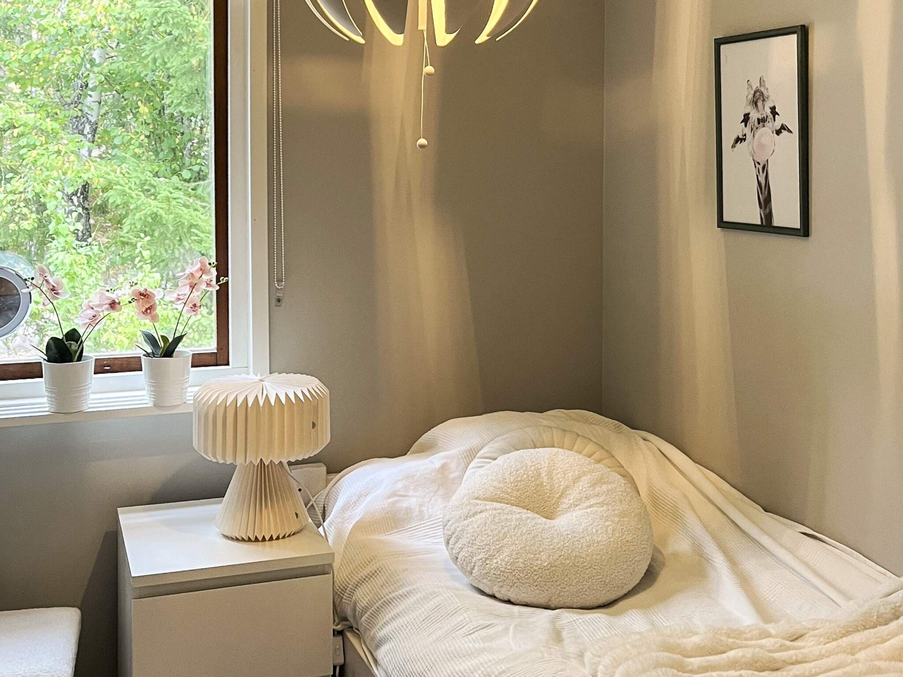 A bedroom with a bed, a bedside table, a lamp, a window with plants, and a framed picture on the wall.