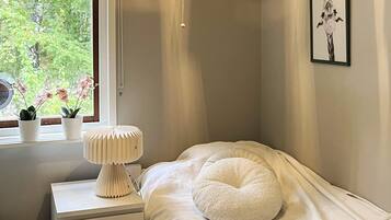 A bedroom with a bed, a bedside table, a lamp, a window with plants, and a framed picture on the wall.