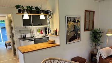 A modern kitchen with a wooden countertop, hanging plants, and a dining area with a wicker chair and a brown ottoman.