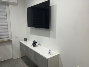 Une pièce avec une console blanche, un téléviseur à écran plat fixé au-dessus, et une fenêtre avec des stores.