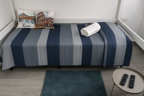 Un lit simple avec une couvre-lit à rayures, un oreiller orné d’un motif sur le thème de Rome et une serviette enroulée posée sur le lit. On aperçoit une petite table ronde avec deux télécommandes posées sur le sol.