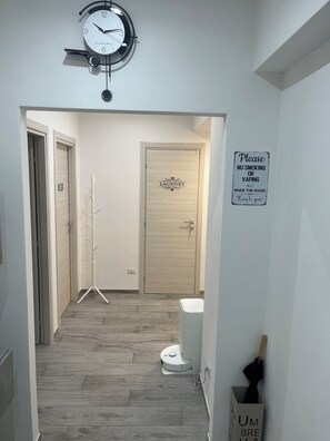 A hallway with a clock, a laundry sign, a no smoking or vaping sign, a coat rack, and a vacuum cleaner. - Domus Acquedotti (Roma)