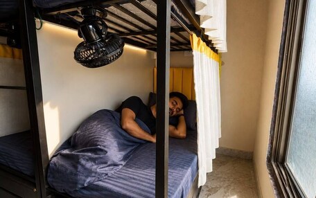 A bunk bed room with a person lying on the lower bunk, a ceiling fan, and a window with curtains.