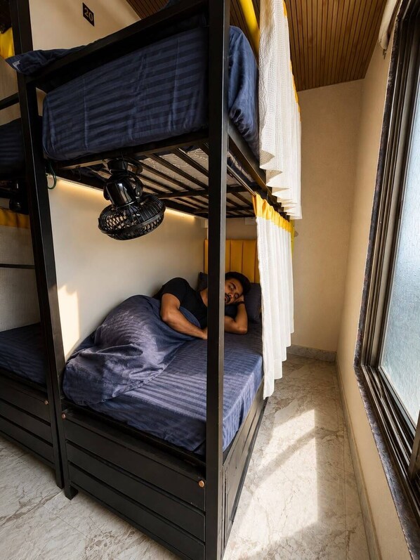 A bunk bed room with a person lying on the lower bunk, a ceiling fan, and a window with curtains.