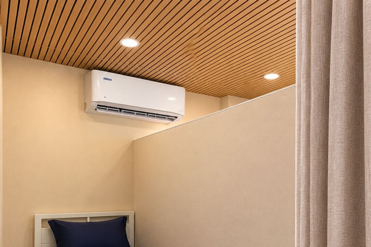 A single bed with a white frame and dark blue bedding, a white wall, a wooden ceiling, and a ceiling-mounted air conditioning unit.
