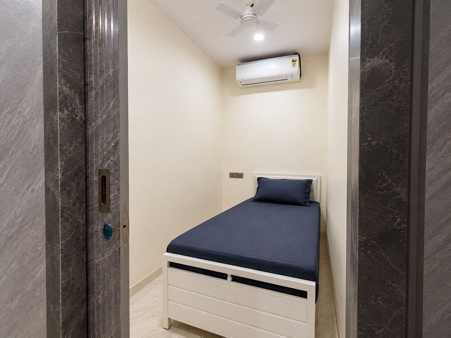 Una stanza piccola e stretta con un letto, una ventola a soffitto e un condizionatore.