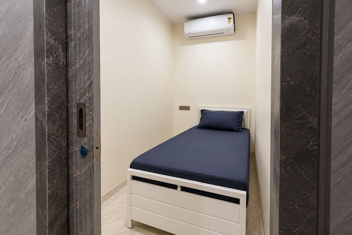 A small, narrow room with a bed, a ceiling fan, and an air conditioning unit.