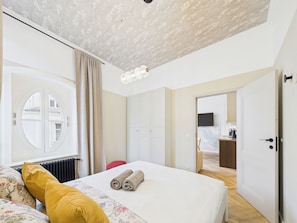 Un dormitorio con cama, ventana con cortinas, radiador y puerta que da a otra habitación.