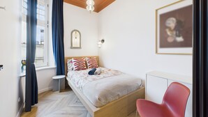 Une chambre à coucher comprenant un lit, une chaise, un bureau, une fenêtre avec des rideaux et un luminaire fixé au mur.