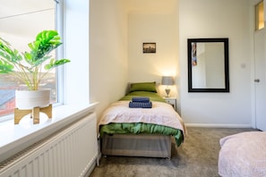 Een slaapkamer met een bed, een nachtkastje met een lamp, een plant in pot en een raam.