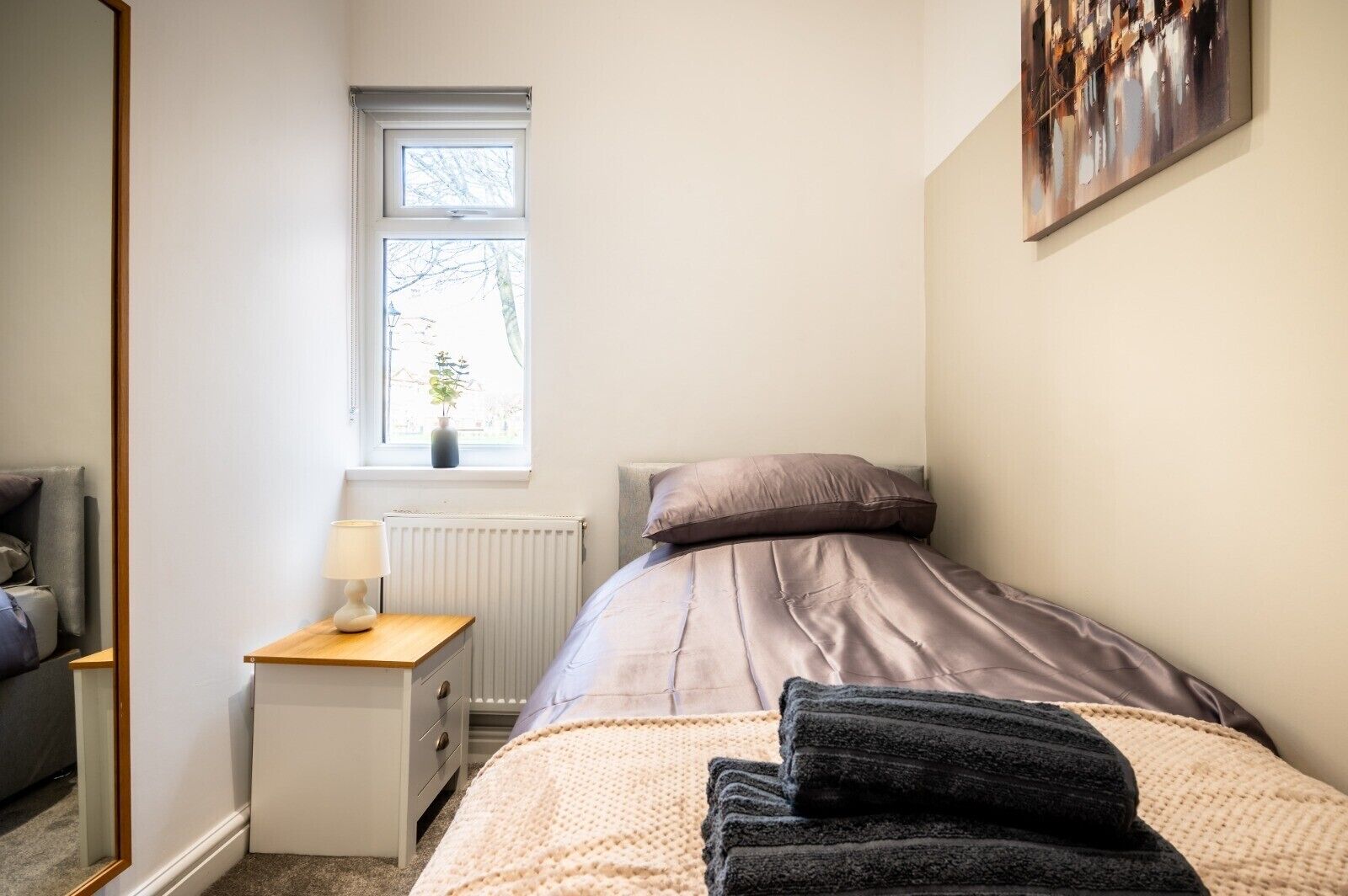 A bedroom with a bed, a nightstand, a lamp, a radiator, a window, and a painting on the wall.