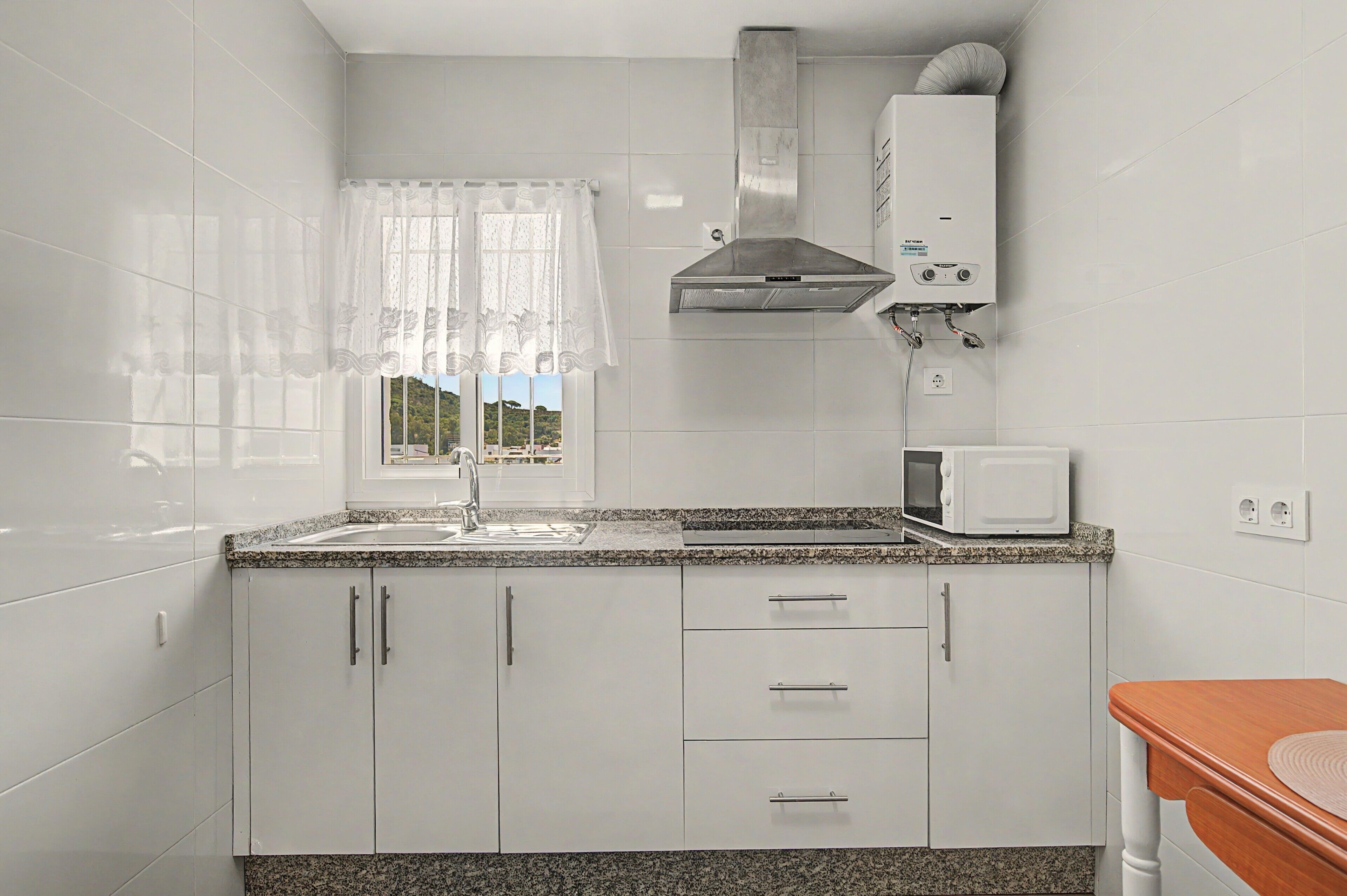 A kitchen with white cabinets, a granite countertop, a window with a white curtain, a stainless steel range hood, and a microwave.