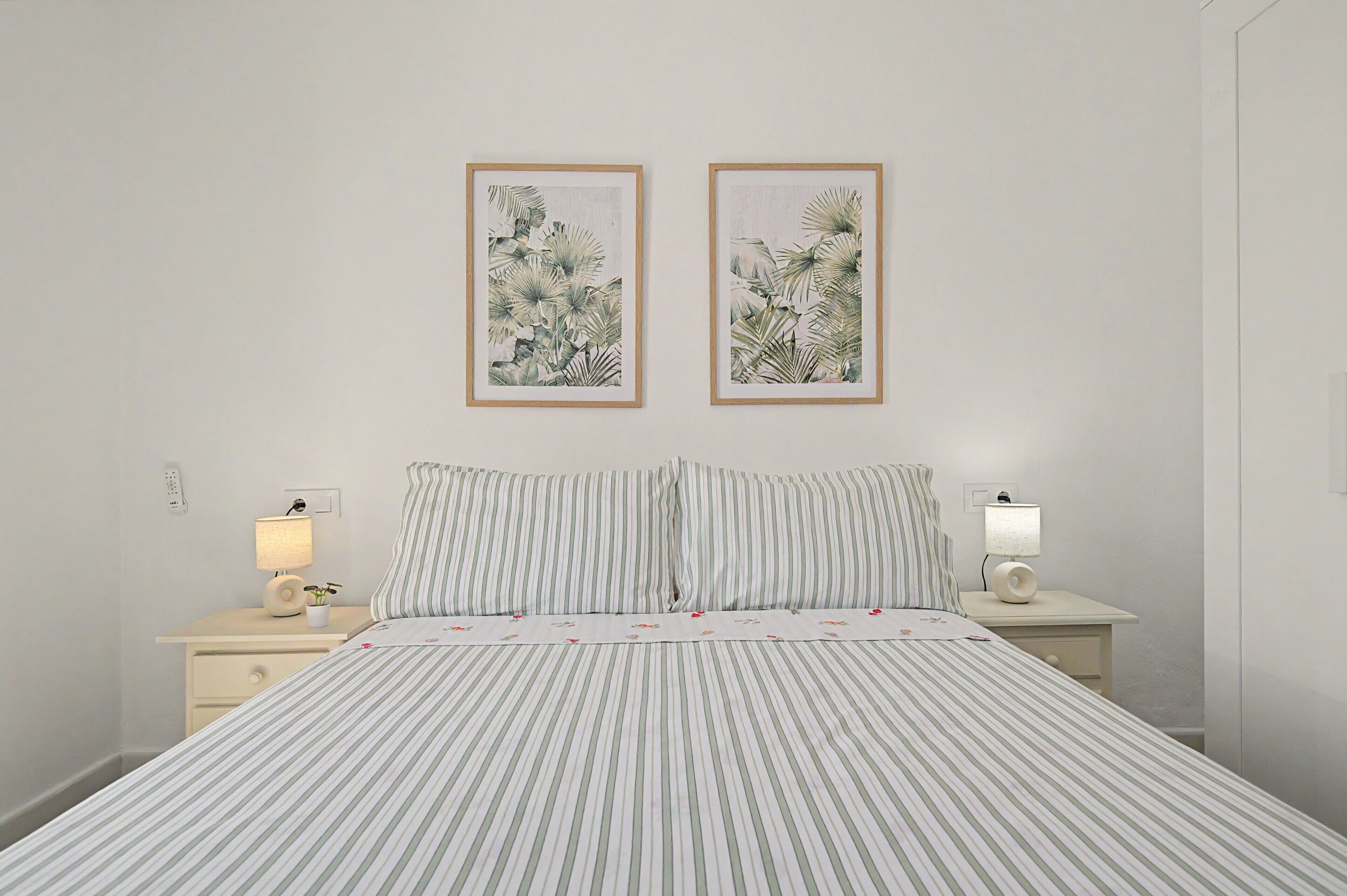 A hotel room with a bed, two nightstands, a lamp, a power outlet, and two framed botanical prints on the wall.