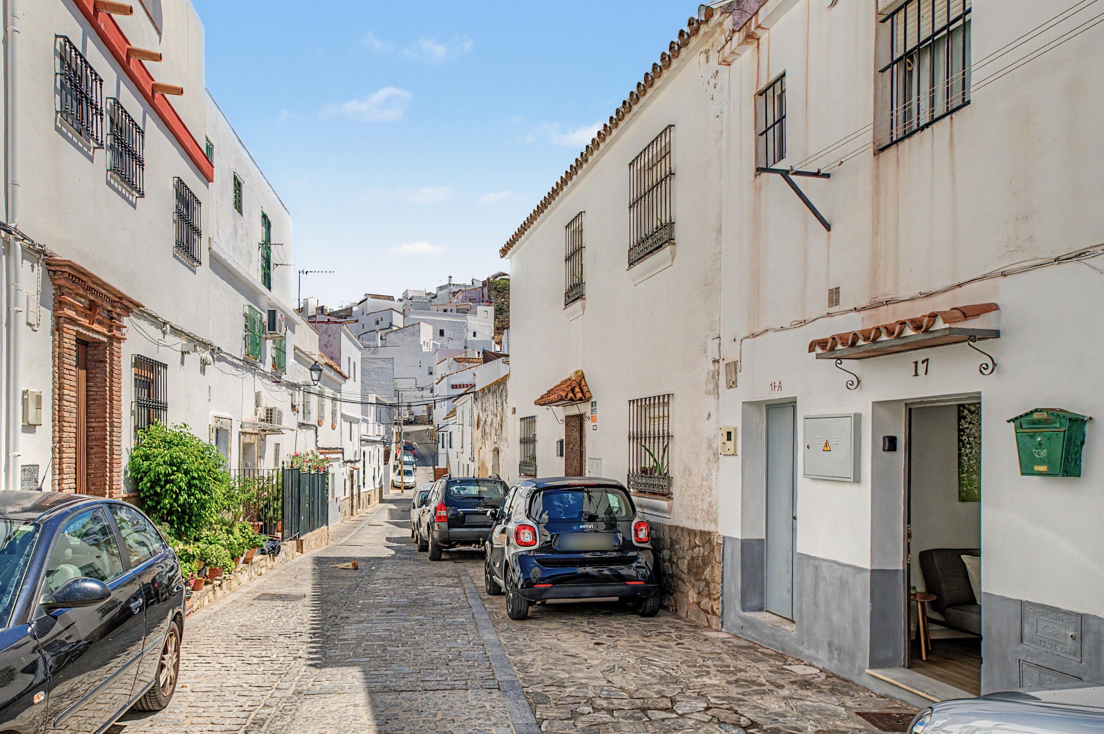 Una estrecha calle empedrada flanqueada por edificios blancos, coches aparcados y una puerta con el rótulo '11A'.