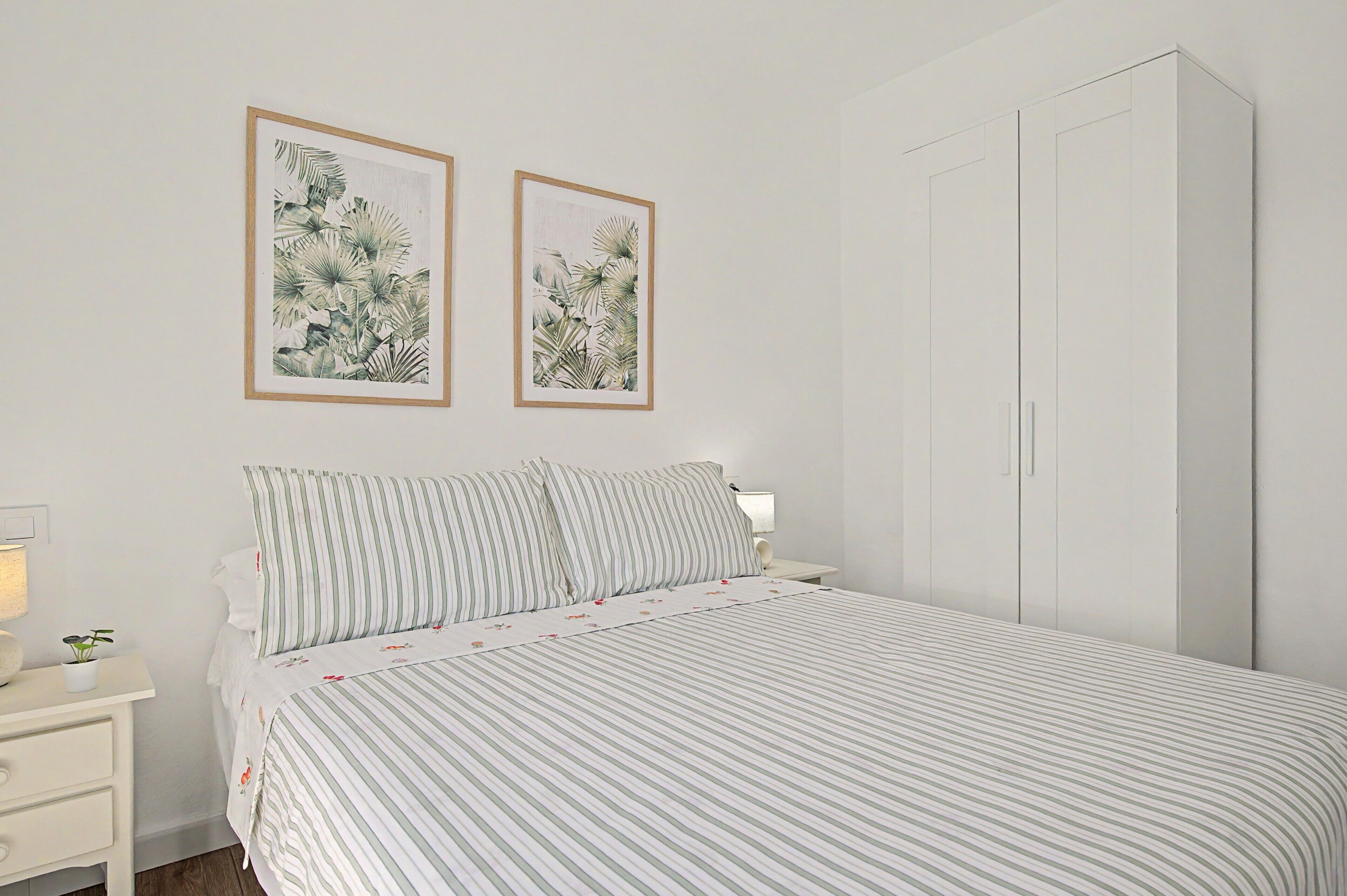 A bedroom with a bed, two framed botanical prints, a bedside table with a lamp, and a wardrobe.
