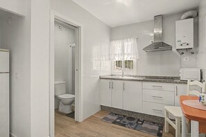 A compact kitchen with white cabinets, a stainless steel range hood, a sink, and a window with lace curtains. - El Rincón de la Mancebía Apartment (Alcalá de los Gazules)
