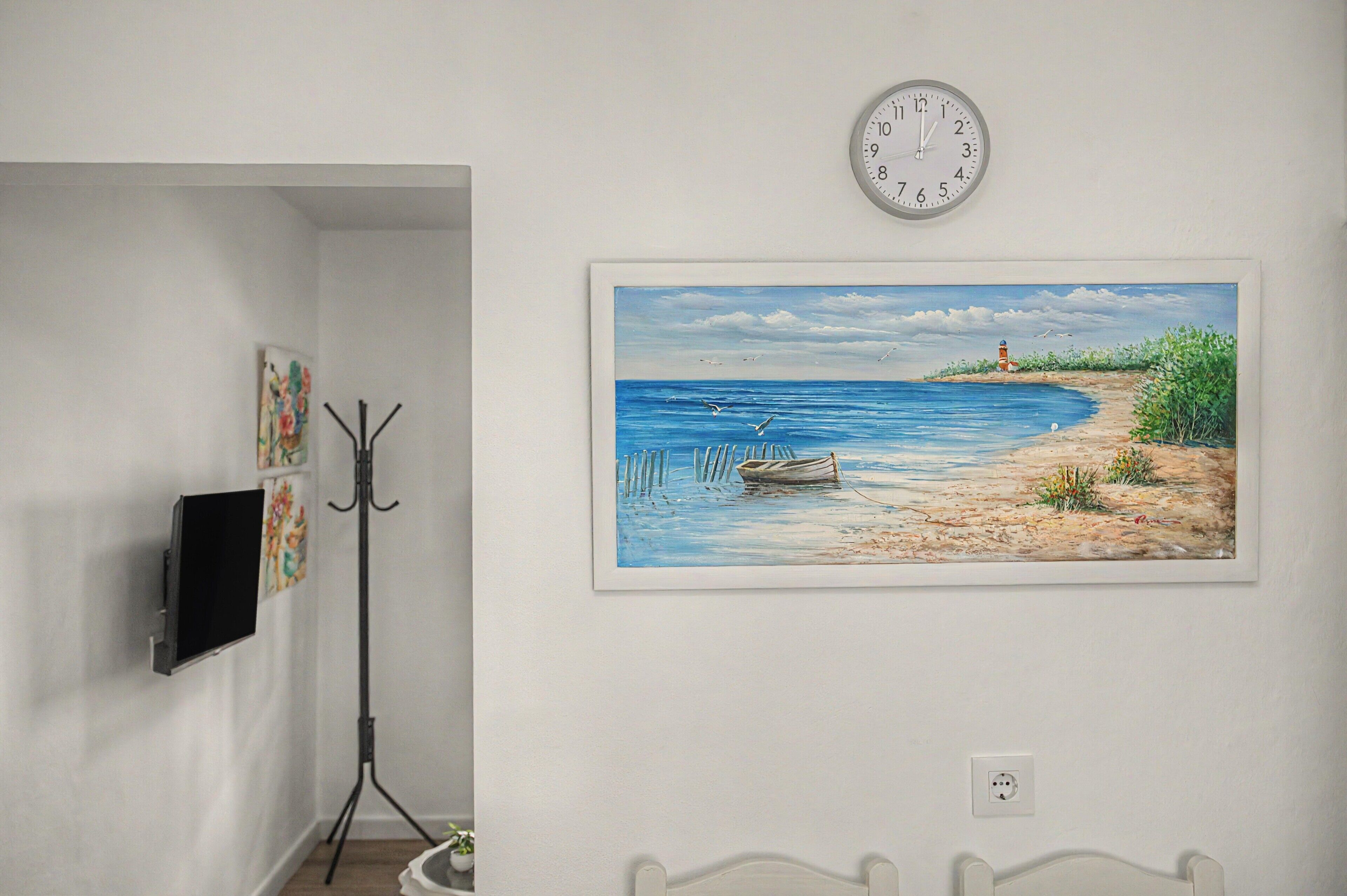 A room with a wall-mounted clock, a framed painting of a beach scene, and a television on a stand.