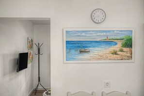 Una habitación con un reloj de pared, un cuadro enmarcados con escena de playa y un televisor sobre un soporte.