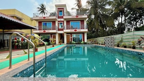 A swimming pool with a two-story building in the background, surrounded by tropical vegetation. - Coral Cove Resort (Alibag)
