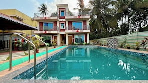 Una piscina con un edificio de dos plantas al fondo, rodeada de vegetación tropical.