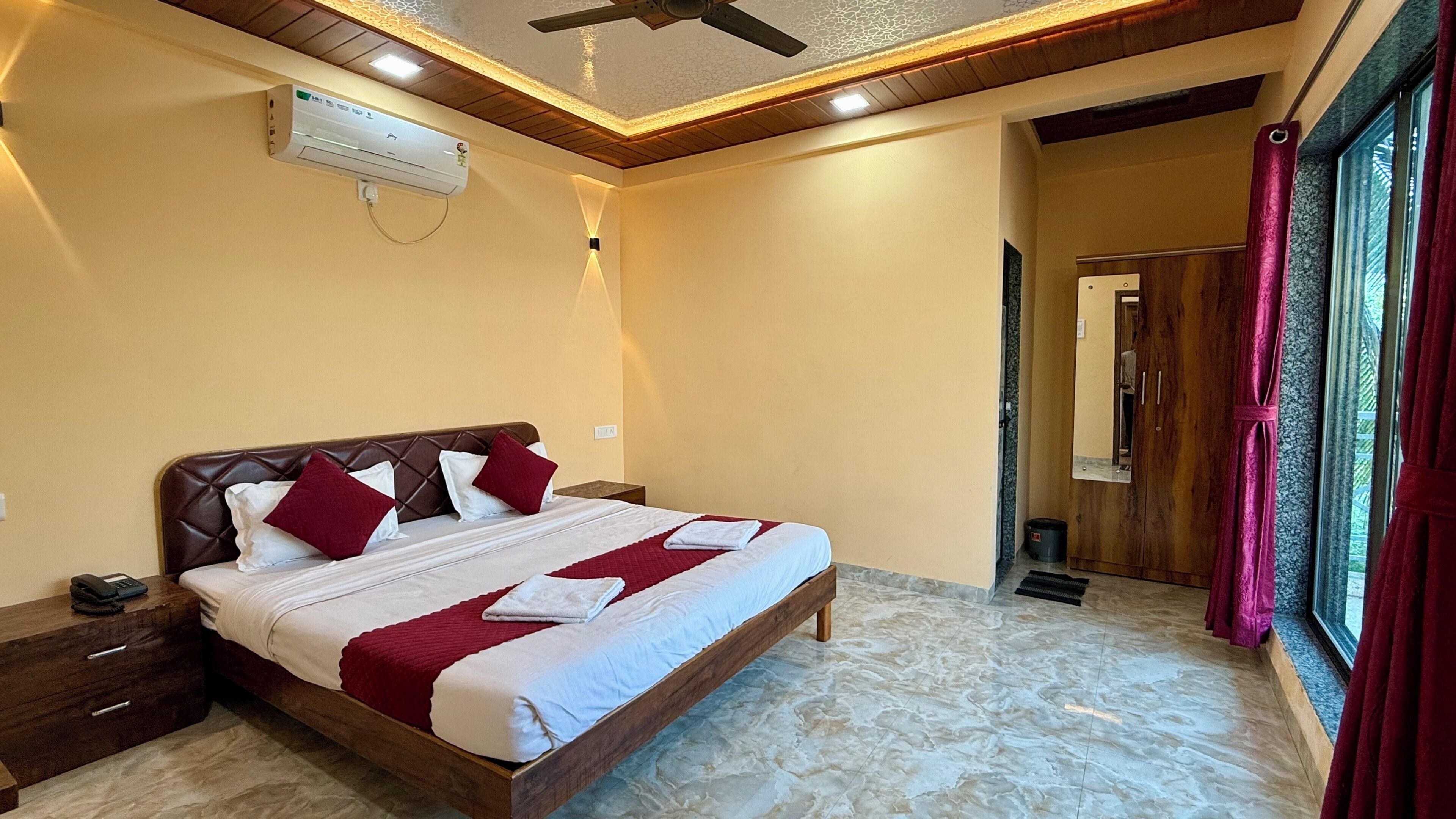 A hotel room with a bed, bedside tables, a ceiling fan, an air conditioning unit, and a window with curtains.