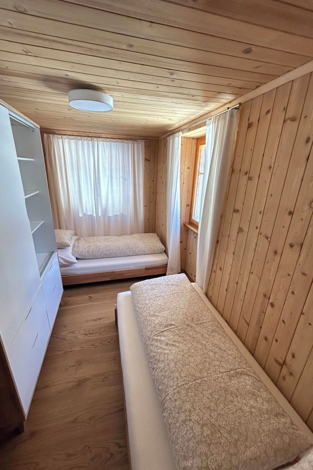 Een knus kamertje met twee bedden, een raam met gordijnen en een plafondlamp.