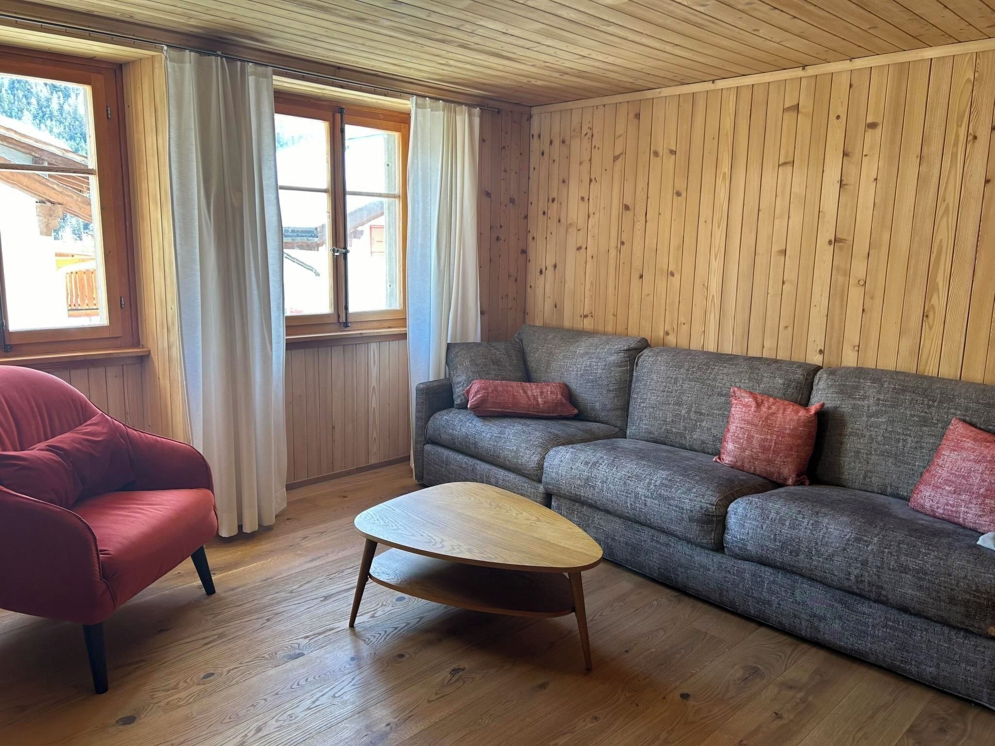 Una stanza con soffitto in legno, un divano grigio con cuscini rossi, una poltrona rossa e un tavolino da caffè in legno.