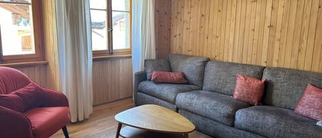 A room with a wooden ceiling, a grey sofa with red cushions, a red armchair, and a wooden coffee table.