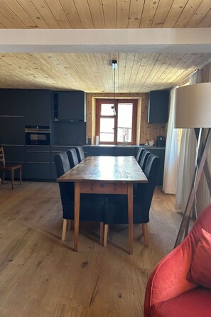 Ein Essbereich mit Holztisch und Stühlen, eine Küche mit dunklen Schränken und ein Fenster mit Tageslicht.
