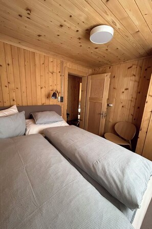 Puisesta hirrestä rakennettu mökki, jossa on kaksi sänkyä, tuoli, lamppu ja ovi.