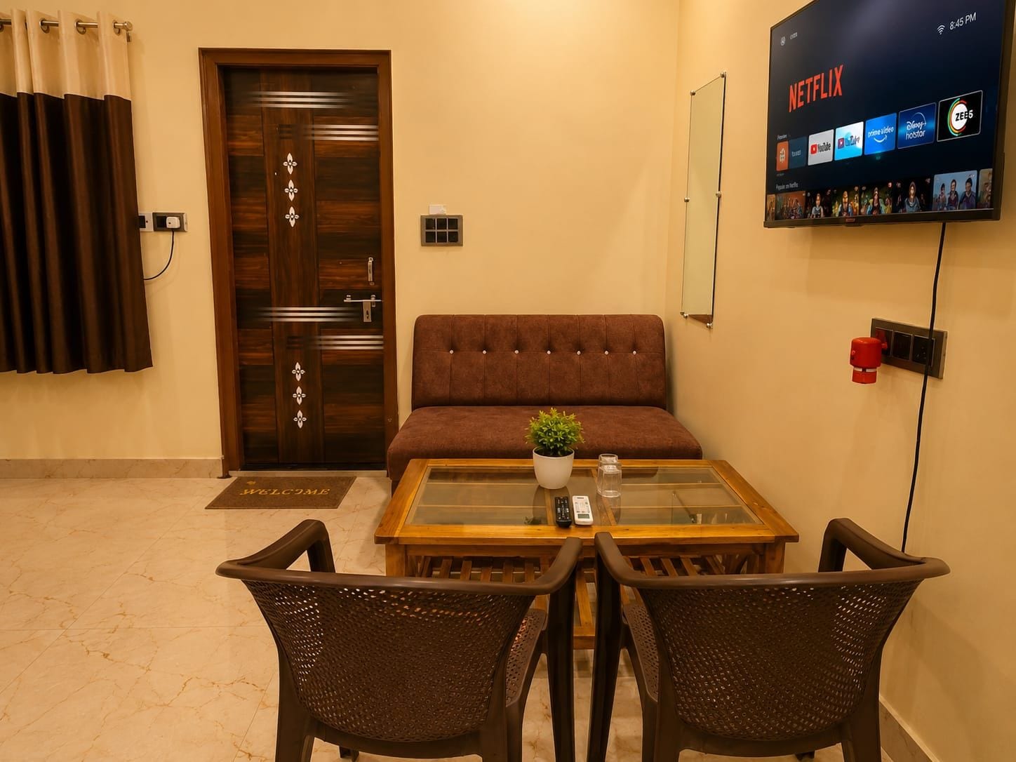 A room with a sofa, a coffee table with a plant, two chairs, a TV displaying Netflix, and a wooden door.