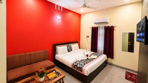 Habitación de hotel con una pared de acento roja, una cama con colcha estampada, un sofá marrón, una mesa de centro de madera con un jarrón de flores, un equipo de aire acondicionado y un televisor en la pared.