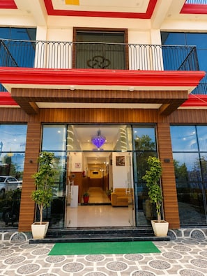 Entrada de un hotel con fachada de vidrio, un toldo rojo y blanco, y un balcón con barandillas de metal.